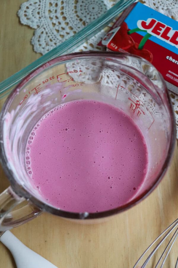 A glass measuring cup filled with a frothy pink liquid sits on a wooden surface beside a box of cherry Jell-O, a whisk, and part of a white lace doily.