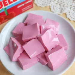 A white plate filled with pink, square-shaped Jello cubes sits on a wooden surface. In the background, a box of cherry-flavored Jell-O and a lacy white cloth are partially visible.