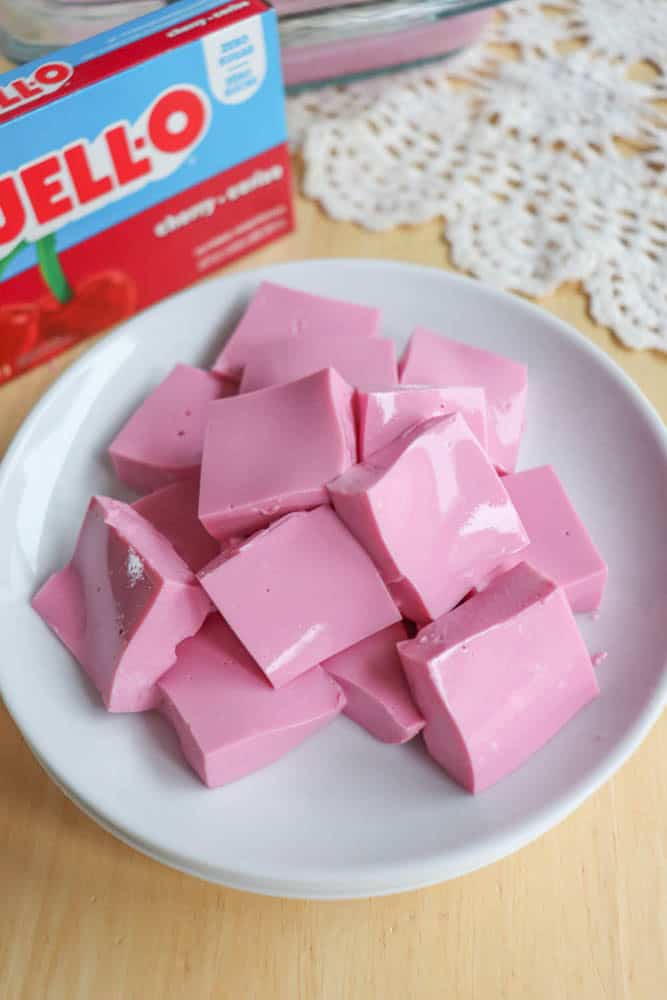 A white plate filled with pink, square-shaped Jello cubes sits on a wooden surface. In the background, a box of cherry-flavored Jell-O and a lacy white cloth are partially visible.