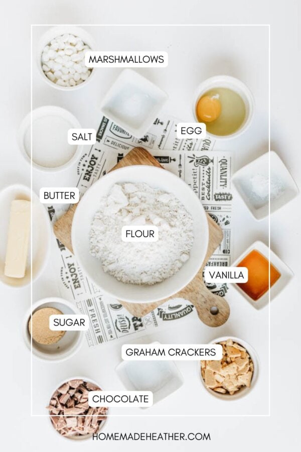 A flat lay of labeled baking ingredients: marshmallows, salt, egg, butter, flour, vanilla, sugar, graham crackers, and chocolate, all arranged around a bowl of flour on a white surface.