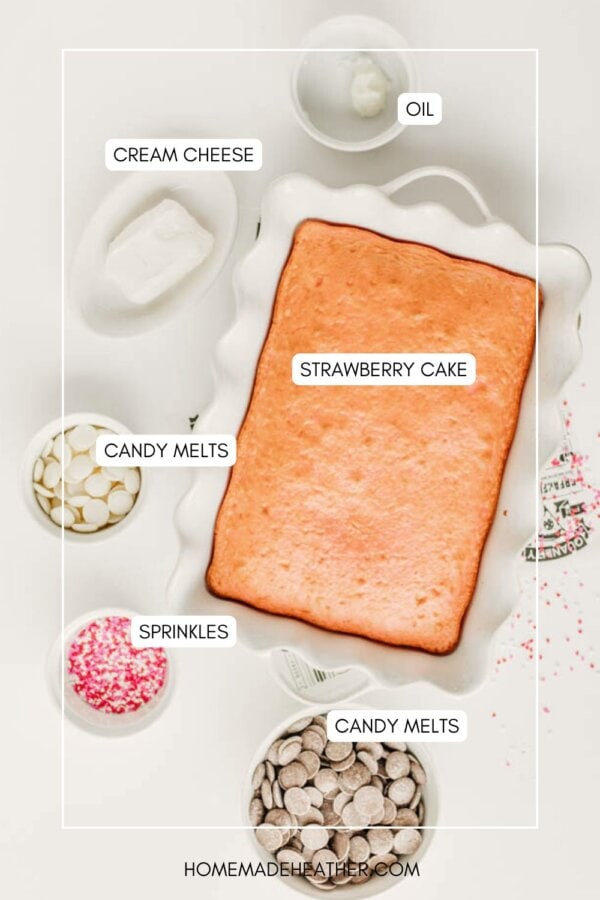 A baked rectangular strawberry cake in a white dish, surrounded by small bowls of cream cheese, oil, pink and white sprinkles, and white and milk chocolate candy melts. Ingredients are labeled.