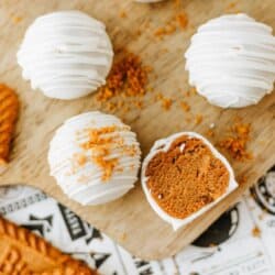 No-bake Biscoff truffles coated in white chocolate are arranged on a wooden board with Biscoff cookies and crumbs scattered nearby. One truffle is halved, revealing its biscoff cookie butter delight filling. Text reads, No-bake Biscoff truffles.
