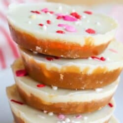 A stack of round Valentines Day cookies with a white chocolate topping, decorated with red, pink, and white heart-shaped sprinkles, on a white plate. Banner text reads Valentines Day Crack Cookies.