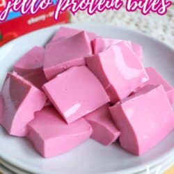 A plate of pink Jello protein bites cut into squares, stacked on white plates. Text on the image reads, 2-ingredient Jello protein bites, so easy. Website URL: homemadeheather.com.