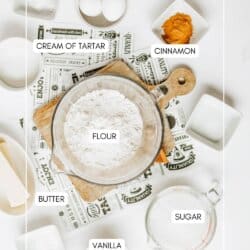 A flat lay of baking ingredients labeled: eggs, cream of tartar, cinnamon, flour, butter, sugar, and vanilla, arranged around a bowl of flour on a wooden board. Recipe cards and small dishes are also visible.