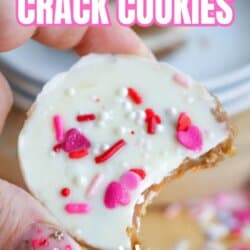 A hand with pink, heart-decorated nails holds a round cookie topped with white icing and pink, red, and white sprinkles. The cookie has a bite taken out. Text reads: Valentines Crack Cookies, So Easy.