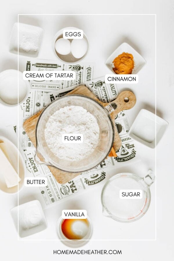 A flat lay of baking ingredients labeled: eggs, cream of tartar, cinnamon, flour, butter, sugar, and vanilla, arranged around a bowl of flour on a wooden board. Recipe cards and small dishes are also visible.