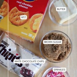 A box of Ritz crackers, a bowl of butter, a bowl of brown sugar, a bag of Hersheyโs white chocolate chips, and a bowl of pink and white sprinkles arranged on a wooden surface.