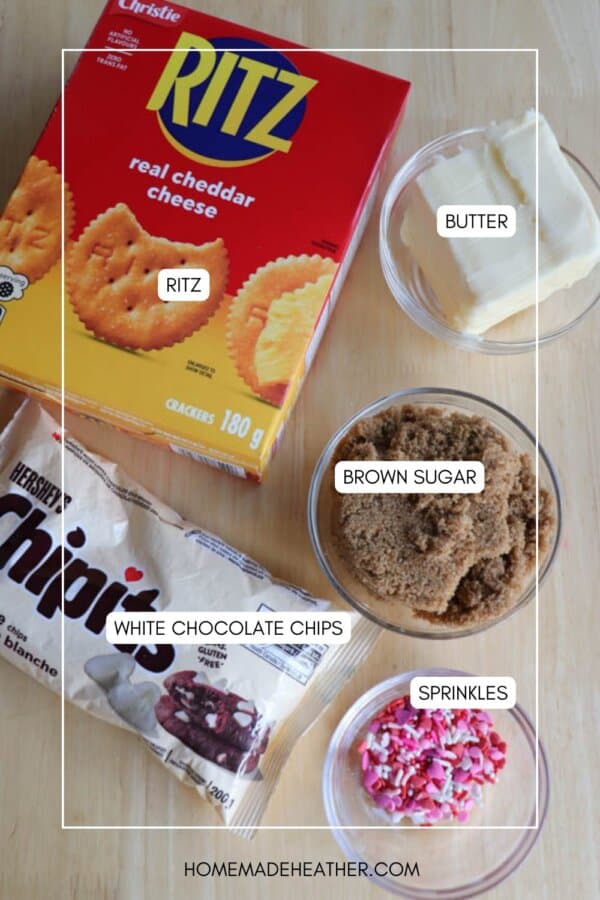 A box of Ritz crackers, a bowl of butter, a bowl of brown sugar, a bag of Hersheyโs white chocolate chips, and a bowl of pink and white sprinkles arranged on a wooden surface.