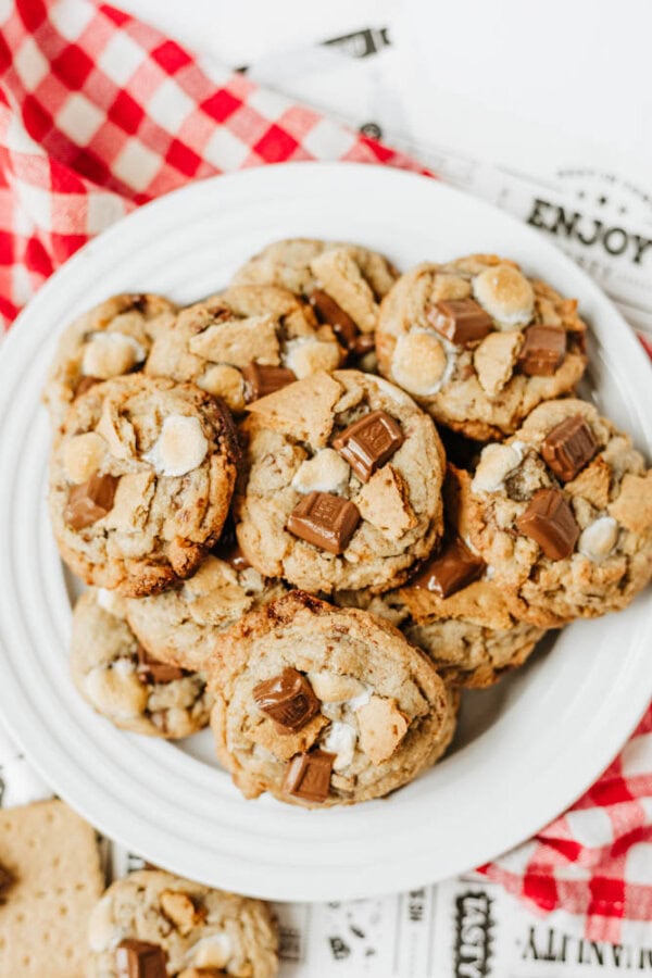 A white plate filled with chocolate chip cookies topped with melted chocolate chunks and marshmallows sits on a red and white checkered cloth.