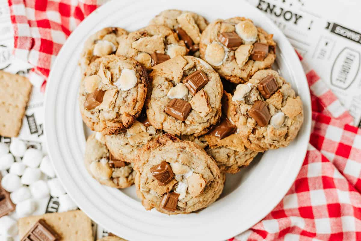 A white plate filled with cookies topped with chunks of chocolate and white marshmallows sits on a red-and-white checkered cloth, surrounded by mini marshmallows, graham crackers, and chocolate pieces.