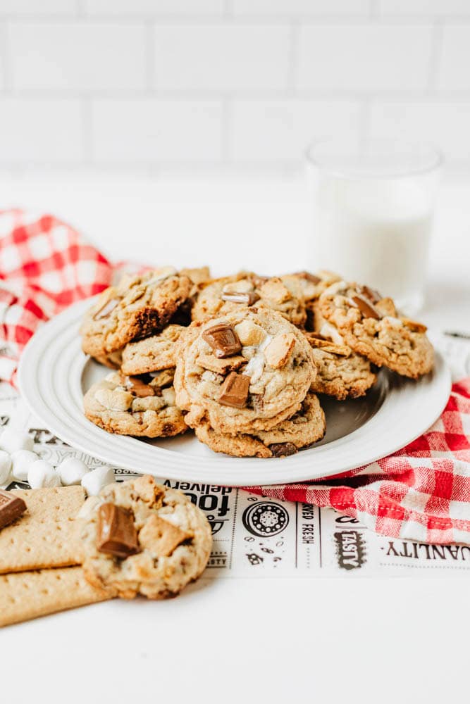 A plate of chocolate chunk cookies on a white table, with a glass of milk in the background and a red-checked napkin underneath. Some cookies and marshmallows are scattered nearby.
