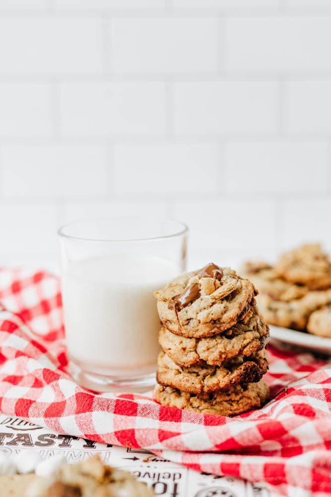 A stack of chocolate chip cookies sits on a red and white checkered cloth next to a glass of milk, with more cookies blurred in the background.