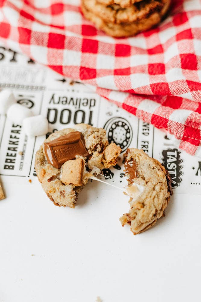 A broken s’mores cookie with melted chocolate, marshmallow, and graham cracker is on a white surface next to a red and white checkered cloth. Marshmallow pieces are scattered nearby.
