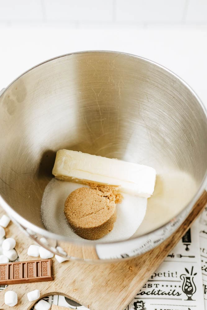 A metal mixing bowl contains a stick of butter, a mound of brown sugar, and a mound of white sugar. The bowl sits on a wooden board with marshmallows and a chocolate bar nearby.