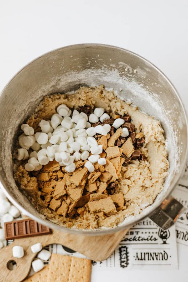 A metal mixing bowl filled with cookie dough, topped with mini marshmallows, chocolate chunks, and pieces of graham crackers, sits on a white surface next to scattered ingredients.