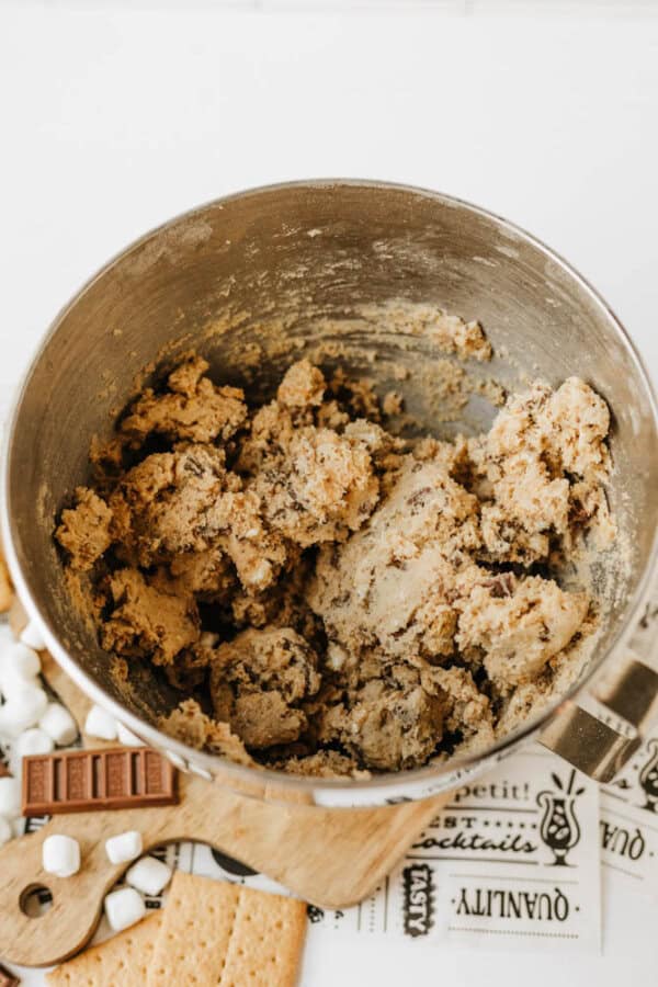 A mixing bowl filled with cookie dough sits on a wooden board surrounded by chocolate pieces, mini marshmallows, and graham crackers on a light surface.