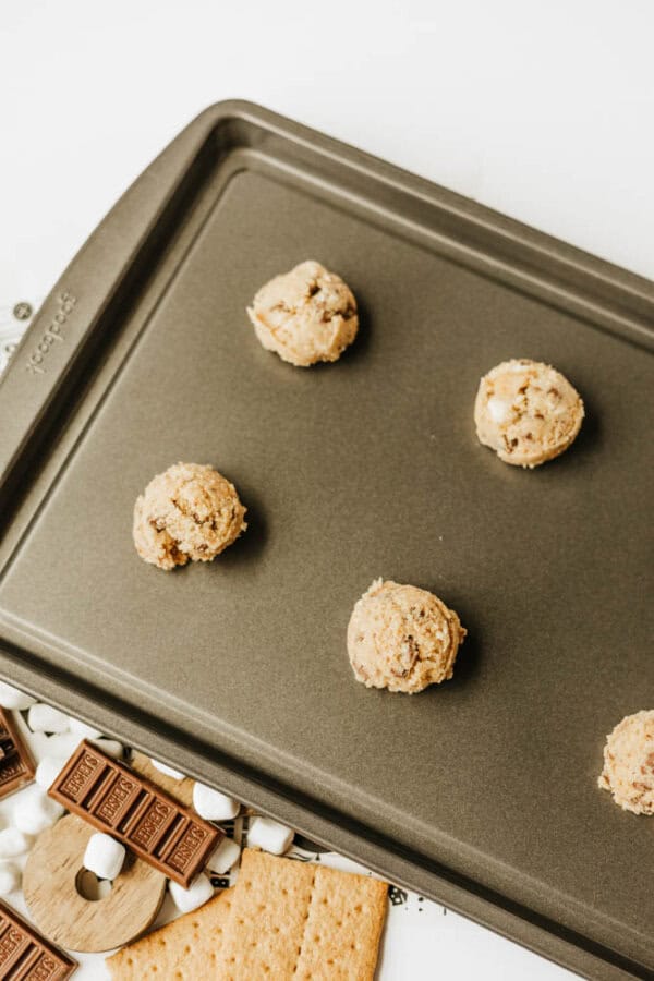 A baking sheet with five scoops of raw cookie dough, placed apart, ready for baking. Nearby are graham crackers, chocolate bars, and mini marshmallows on a white surface.