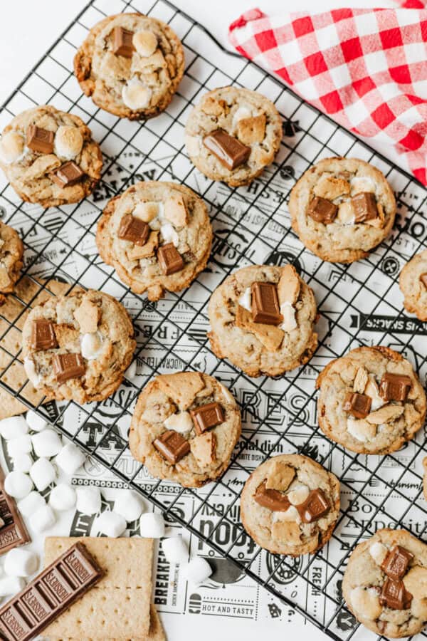 A cooling rack holds several smores cookies topped with chocolate pieces and mini marshmallows. A red and white checkered cloth, chocolate bars, mini marshmallows, and graham crackers are nearby.
