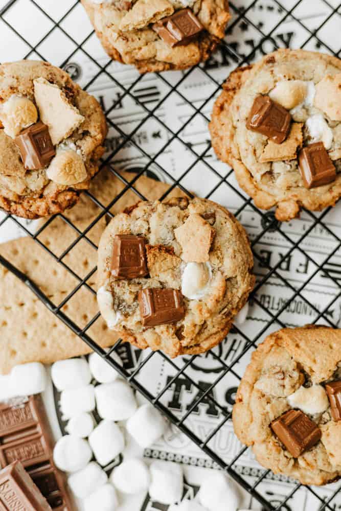 Cookies topped with chocolate chunks, marshmallows, and graham cracker pieces are cooling on a wire rack. Below, there are mini marshmallows, chocolate squares, and part of a graham cracker.