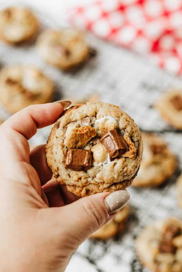 A hand with manicured nails holds a chunky cookie topped with pieces of chocolate and marshmallow, with more cookies on a cooling rack in the blurred background.