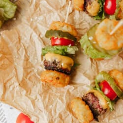 Mini burger skewers with tater tots, lettuce, tomato, pickle, and melted cheese are arranged on crumpled parchment paper, creating a fun and colorful appetizer display.