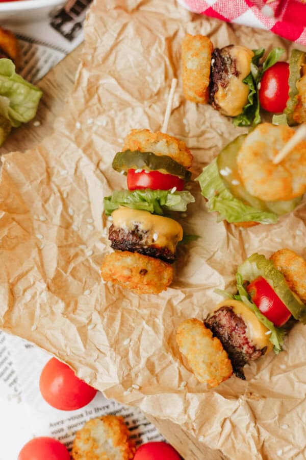 Mini burger skewers with tater tots, lettuce, tomato, pickle, and melted cheese are arranged on crumpled parchment paper, creating a fun and colorful appetizer display.