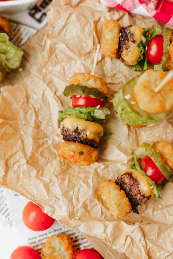 Mini burger skewers with tater tots, lettuce, tomato, pickle, and melted cheese are arranged on crumpled parchment paper, creating a fun and colorful appetizer display.