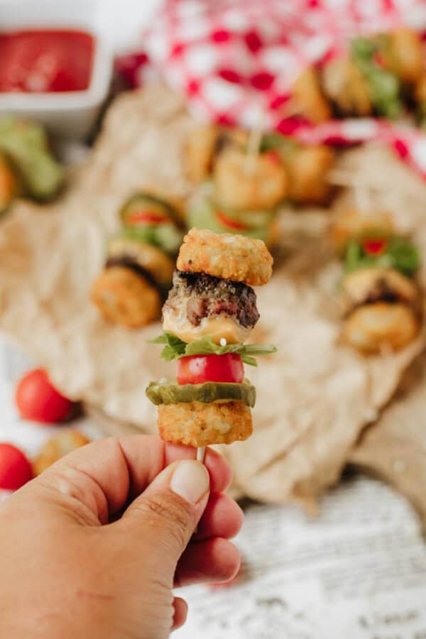 A hand holds a skewer with mini burger ingredients, including a meat patty, lettuce, tomato, pickles, and tater tots, with more skewers and a dish of ketchup in the blurred background.