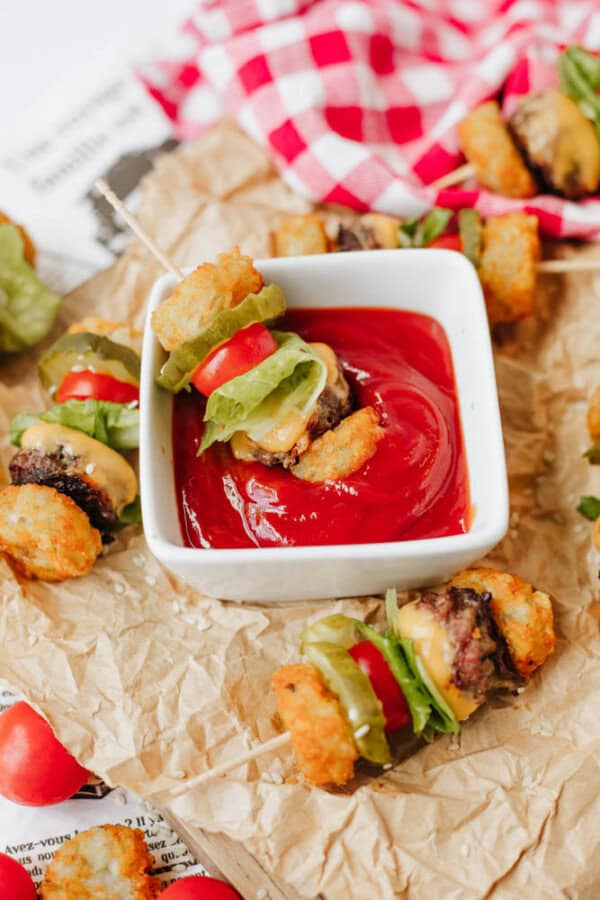 Mini cheeseburger skewers with tater tots, lettuce, pickles, and tomato are arranged around a square bowl of ketchup, with one skewer being dipped. The scene is set on crumpled parchment paper with a red checkered cloth.