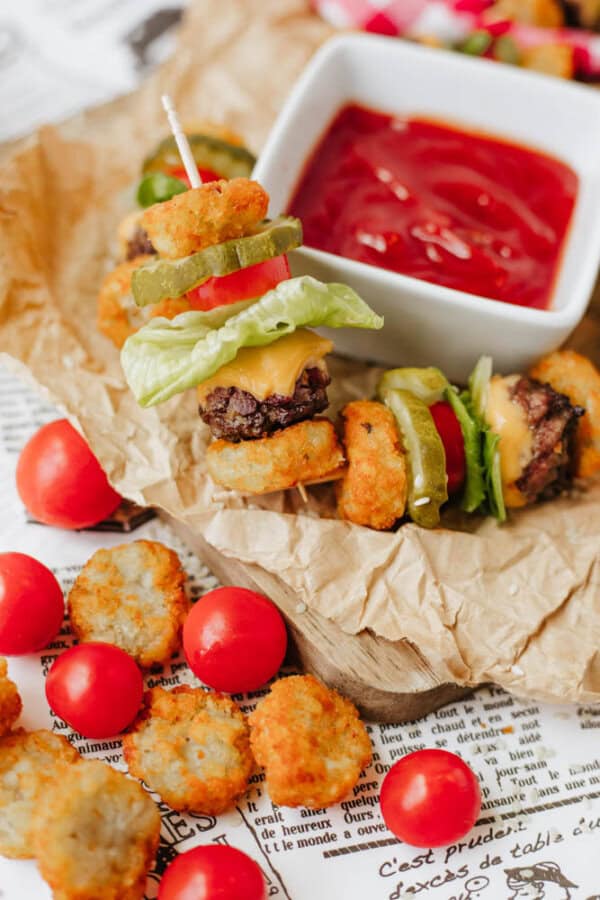 Mini burger skewers with lettuce, cheese, pickles, and tater tots are arranged on parchment paper next to cherry tomatoes and a bowl of ketchup for dipping.