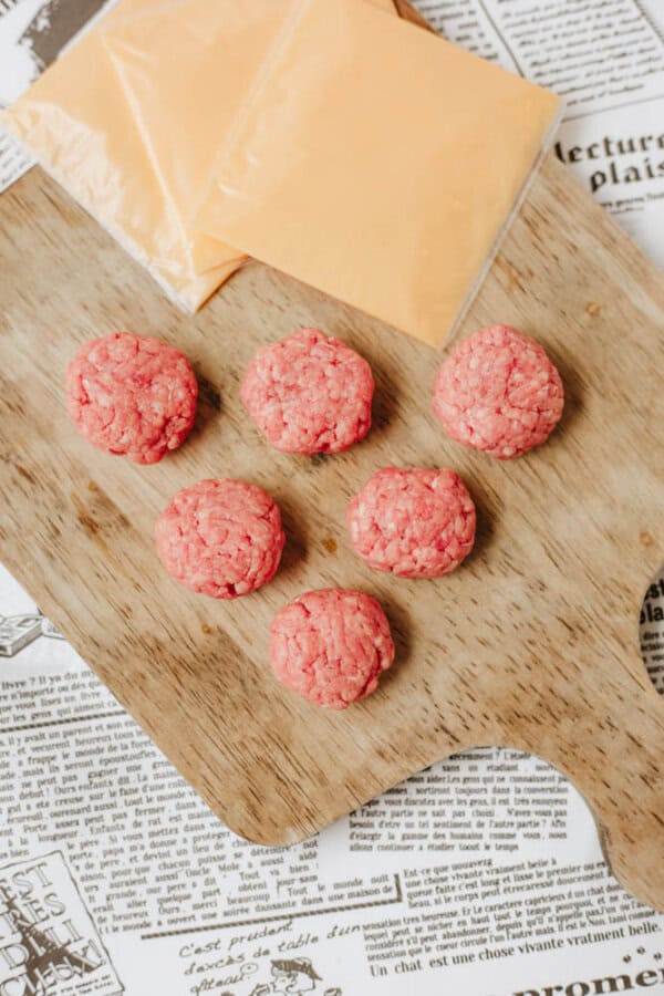 Seven raw ground beef sliders are arranged on a wooden cutting board. Two slices of processed cheese are placed nearby. The background consists of sheets of printed newspaper.