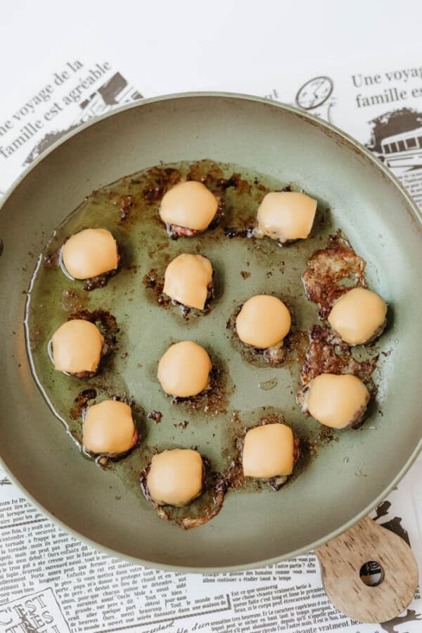 A frying pan with small burger patties topped with melted cheese slices, cooking in oil. The pan rests on sheets of newspaper with French text.