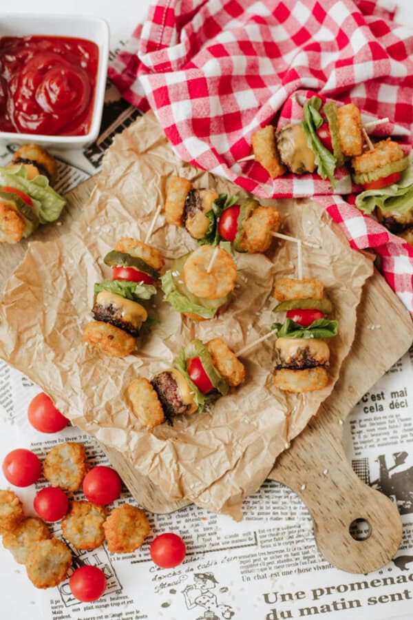 Mini sliders made with tater tots, lettuce, cherry tomatoes, and burger patties on toothpicks are arranged on parchment paper and a wooden board, with ketchup and more tater tots nearby.