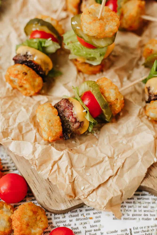Mini burger skewers with tater tots, cheese, pickles, lettuce, cherry tomatoes, and beef patty pieces are arranged on crumpled parchment paper atop a wooden board.