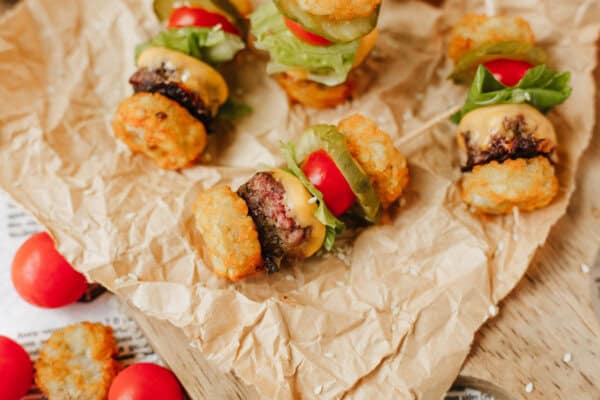Mini sliders made with tater tots as buns, filled with burger patties, melted cheese, lettuce, tomato, and pickles, arranged on crumpled parchment paper on a wooden surface.