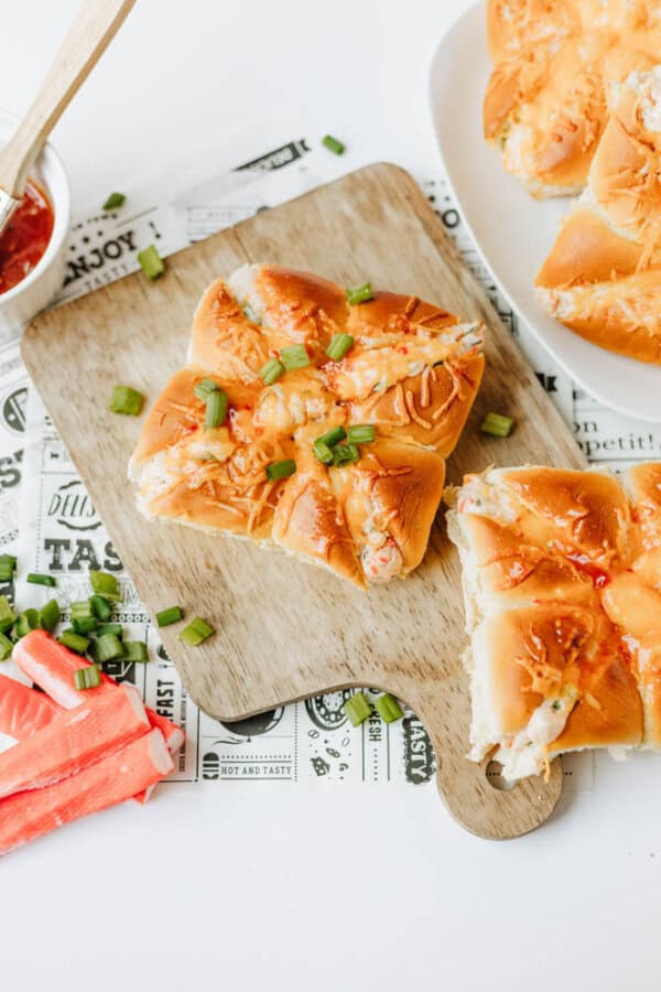 Crab bread rolls topped with chopped green onions on a wooden board, with imitation crab sticks and a dish of tomato sauce nearby. Another plate of rolls is partially visible in the background.