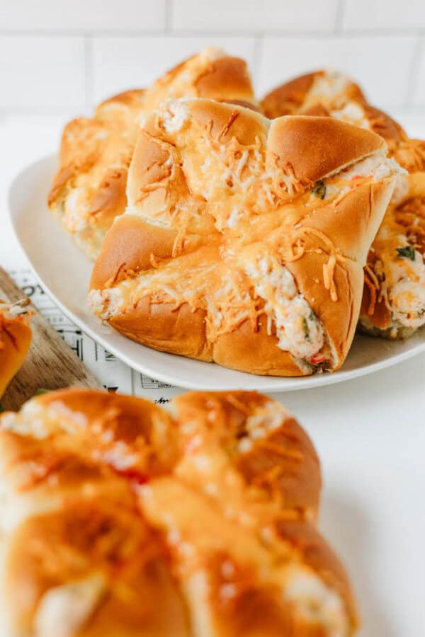 A plate of golden brown bread rolls topped with melted cheese, with more cheese-topped rolls in the foreground. The rolls are fluffy and soft, set on a white surface with a white tiled background.
