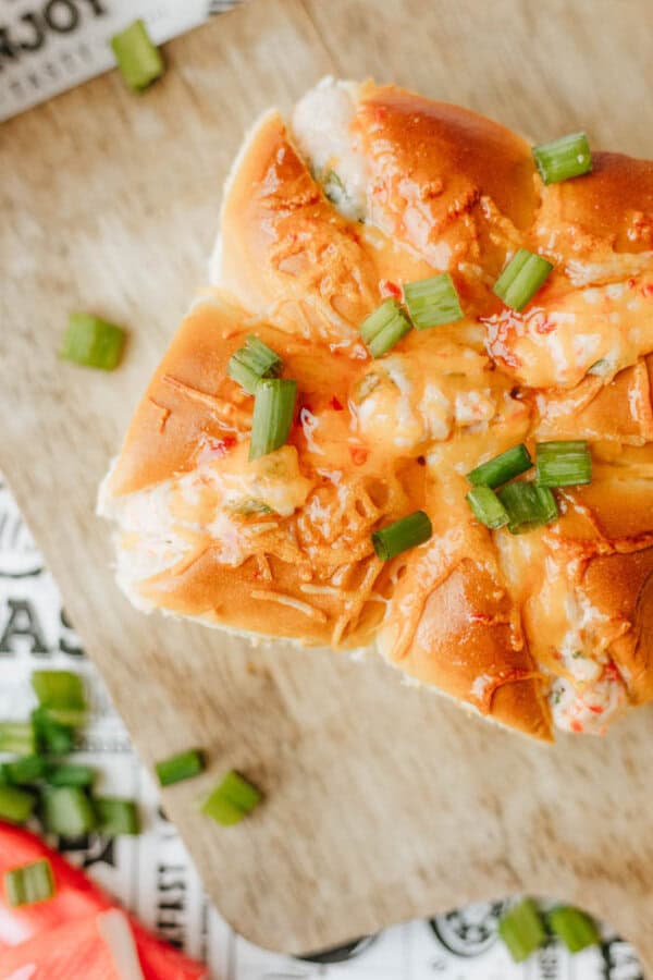 A close-up of a cheesy baked bread roll topped with chopped green onions, served on a wooden board. Some green onion pieces are scattered around the bread.