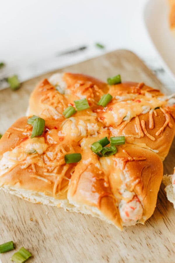 A close-up of a baked bread roll topped with melted cheese, chopped green onions, and a creamy filling, placed on a wooden cutting board.