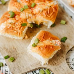 A wooden board with cheesy baked sliders topped with chopped green onions, one slider separated from the rest. The board sits on paper with decorative text and designs.