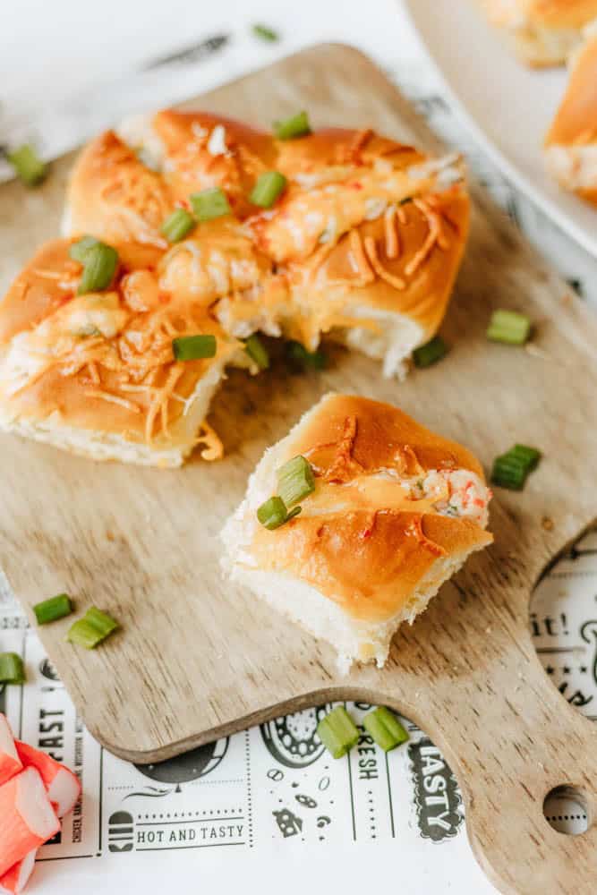 A wooden board with cheesy baked sliders topped with chopped green onions, one slider separated from the rest. The board sits on paper with decorative text and designs.