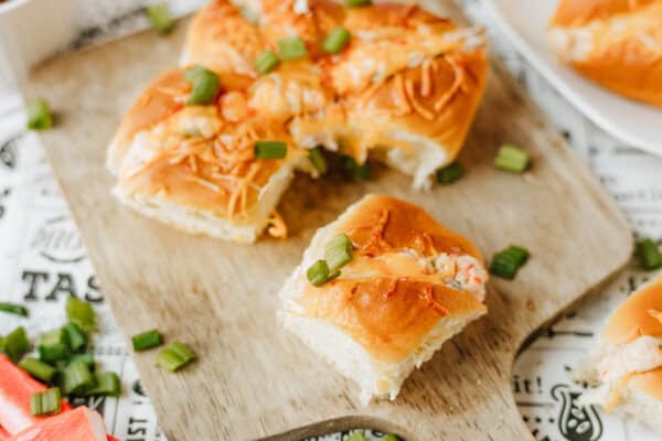 Cheesy baked sliders topped with chopped green onions on a wooden board, with some sliders cut and separated. Green onion pieces are scattered around, and part of a crab stick is visible on the side.