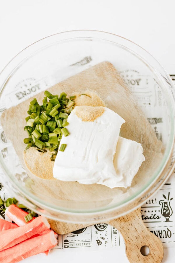 A glass bowl containing cream cheese, chopped green onions, and seasoning sits on a wooden board. Pieces of imitation crab are nearby, and the background features a printed kitchen-themed cloth.