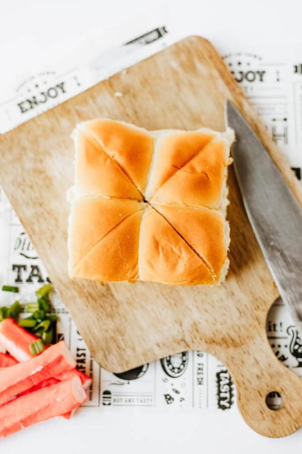 Six soft dinner rolls are placed together on a wooden cutting board beside a knife; imitation crab sticks and chopped green onions sit nearby, all on top of a paper with food-related designs and text.