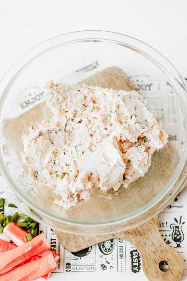 A glass bowl filled with a creamy crab dip mixture sits on a wooden board, with chopped imitation crab and green onions nearby on a white surface.