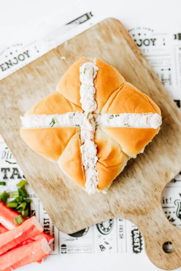 Four dinner rolls arranged in a square on a wooden board, filled in a cross shape with a creamy white salad mixture, likely crab or seafood, garnished with green onions.