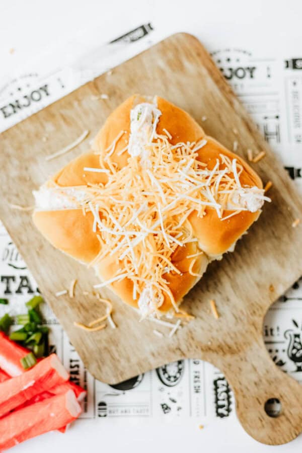 A sandwich with shredded cheese and a creamy filling sits on a wooden cutting board, with sliced green onions and imitation crab pieces nearby. The background includes a paper with food-related text and designs.