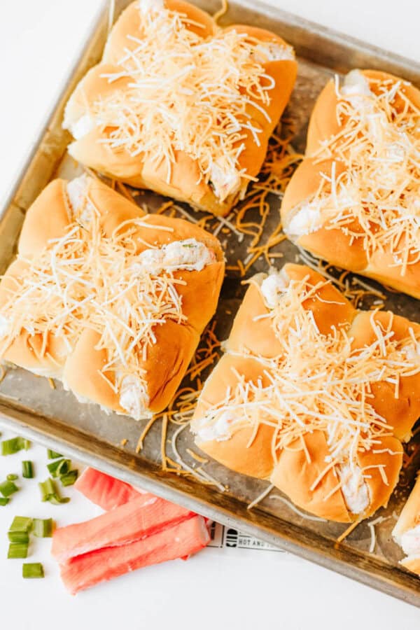 Four sandwich buns filled with a creamy mixture and topped with shredded cheese are arranged on a baking tray. Chopped green onions and imitation crab sticks are placed nearby on the white surface.