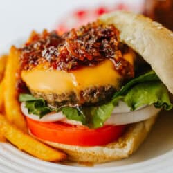 Close-up of a cheeseburger with lettuce, tomato, onion, cheddar cheese, and a generous topping of caramelized onion jam, served on a sesame bun with French fries on a white plate.